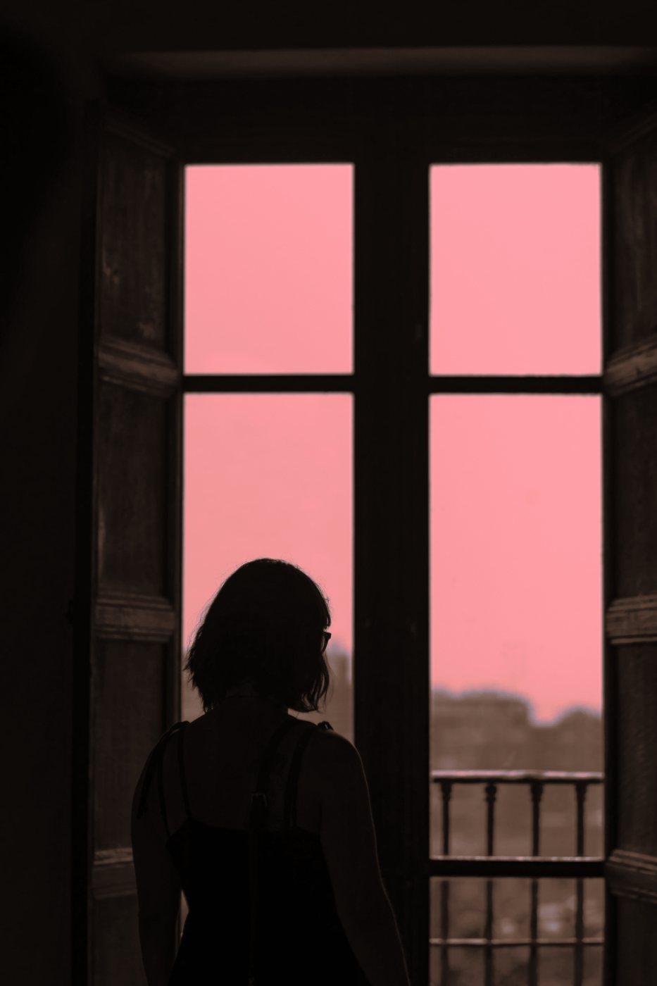 Mulher de costas, em silhueta, diante de uma janela aberta para um céu rosado